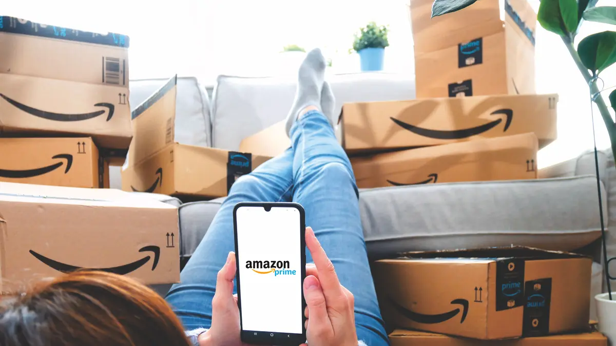 Eine Frau liegt auf dem Sofa mit dem Handy in der Hand, im Hintergrund ein Stapel mit Paketen von Amazon.