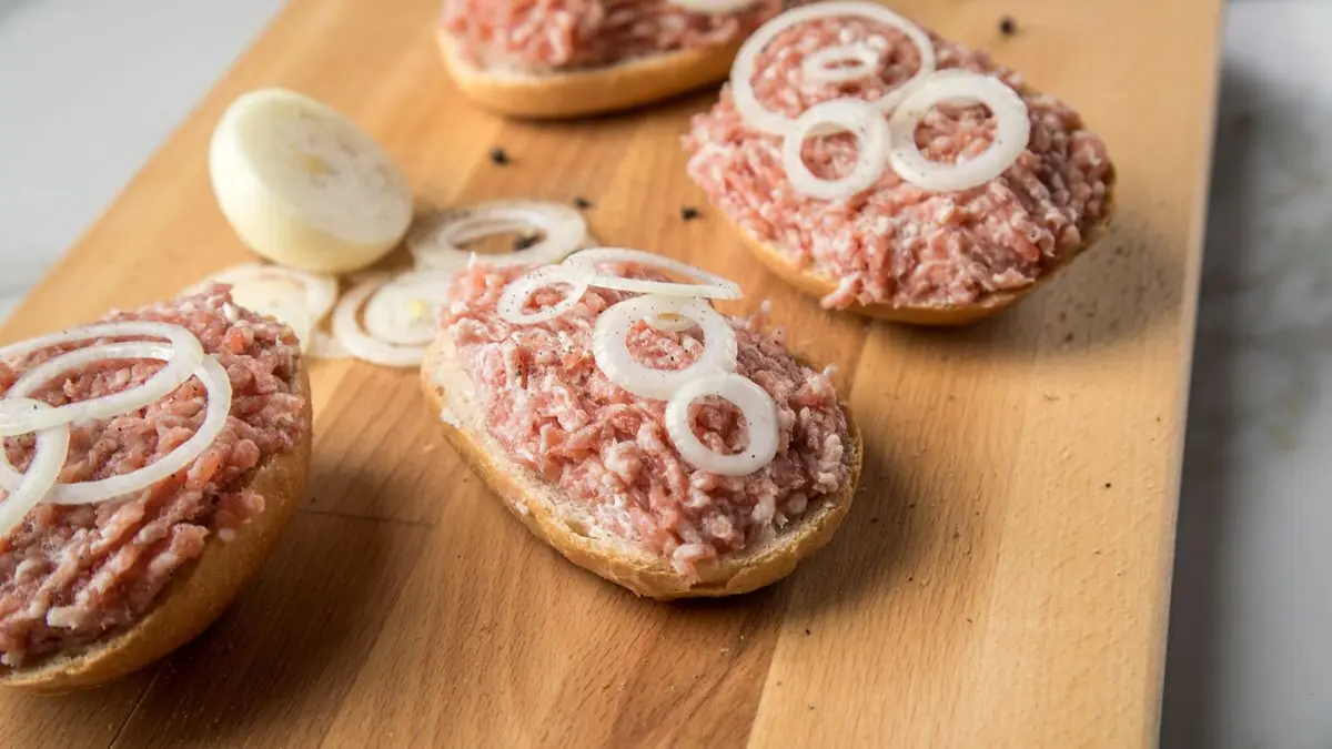 Vier Mettbrötchen mit Zwiebelringen liegen auf einem Holzbrett.