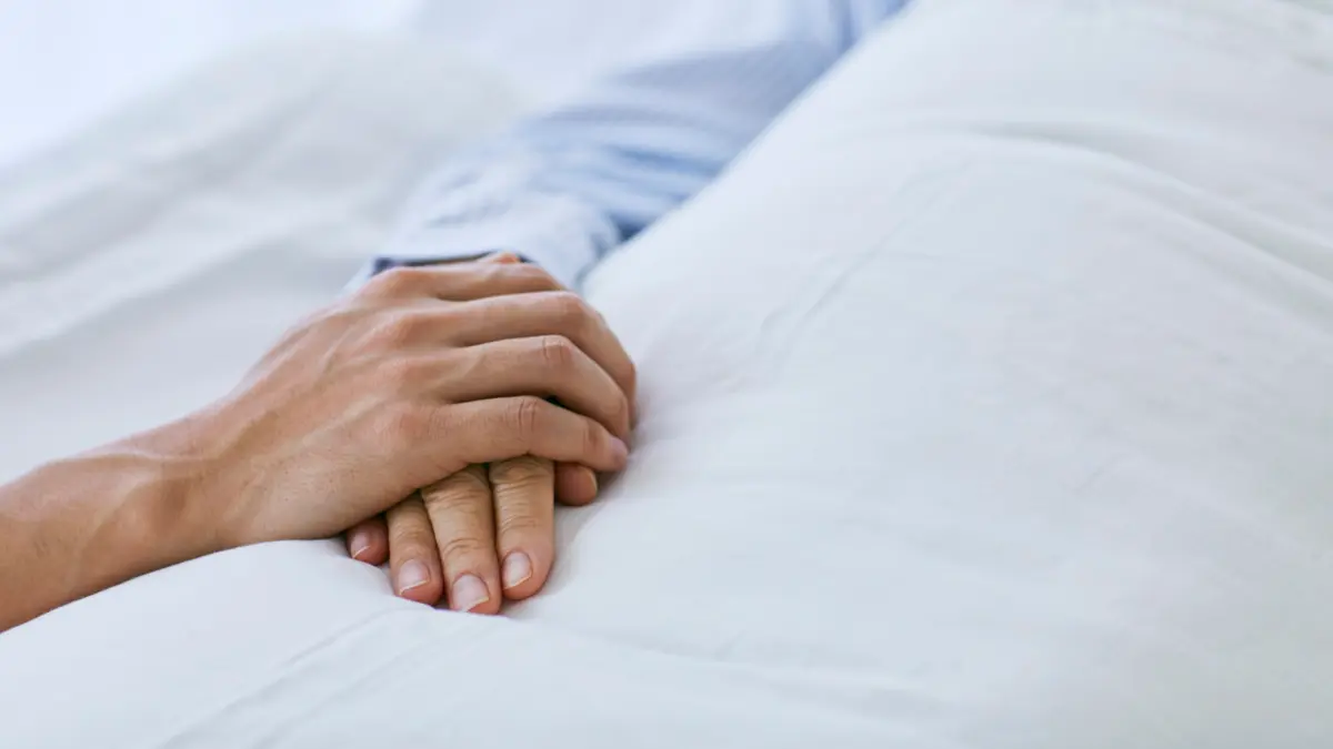 eine Person legt ihre Hand auf die einer im Krankenhausbett liegenden weiteren Person