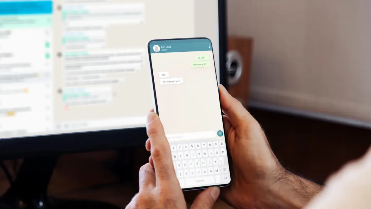 Smartphone in Hand, Messenger geöffnet, im Hintergrund angeschalteter Monitor