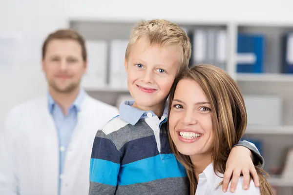 Mutter und Sohn in der Zahnarztpraxis. Sie lächeln in die Kamera, im Hintergrund der Kieferorthopäde