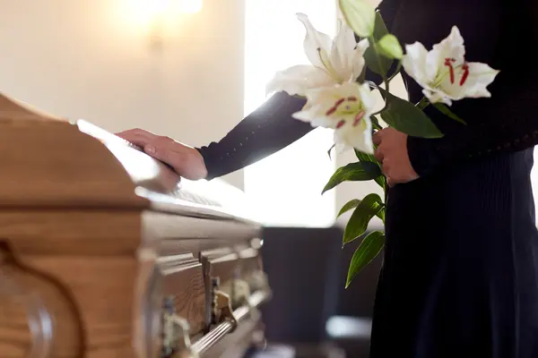 Eine Frau steht mit Blumen in der Hand an einem Sarg.
