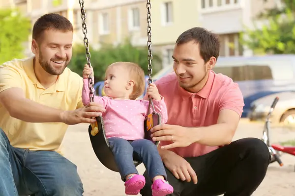Zwei Männer spielen mit ihrer Tochter, die auf einer Schaukel sitzt.