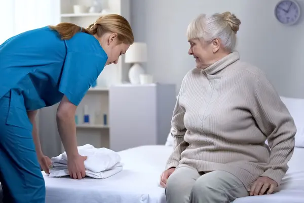 Eine ältere Dame sitz auf ihrem Bett, eine Pflegerin legt Bettwäsche neben ihr ab