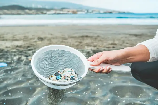 Frau am Strand sammelt mit einem Sieb kleine Plastikteilchen aus dem Sand