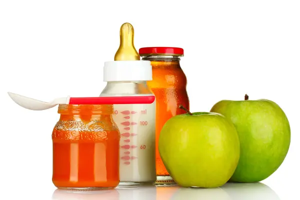 Im Vordergrund links steht ein Gläschen mit orange-rotem Babybrei. Darauf liegt ein Plastiklöffel für Babys. Rechts daneben liegen zwei grüne Äpfel. Im Hintergrund steht eine Babyflasche aus Glas, gefüllt mit Milch und eine kleine, verschlossene Glasflasche mit Apfelsaft.