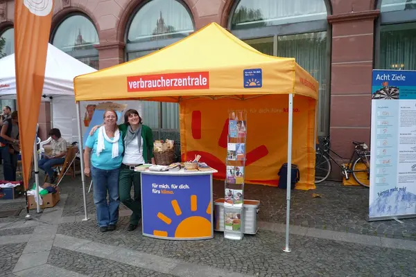 Zwei Frauen stehen an einem Straßenstand mit Infomaterial und blicken in die Kamera.