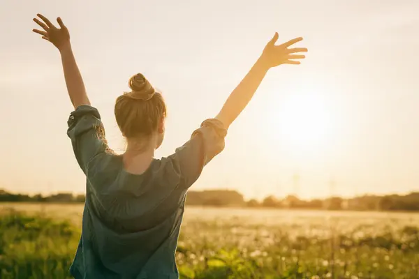 Eine junge Frau steht vor einem Getreidefeld in der Sonne und streckt die Arme in die Höhe.