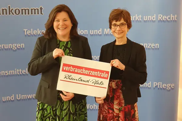 Zwei Frauen stehen vor einer baluen Wand und halten ein Schild mit Verbraucherzentrale Rheinland-Pfalz zwischen sich.