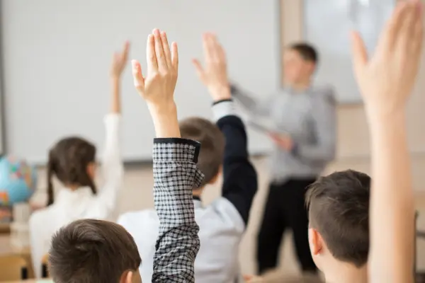 Schulkinder zeigen während einer Unterrichtseinheit im Klassenraum auf, ein Lehrer steht an der Tafel