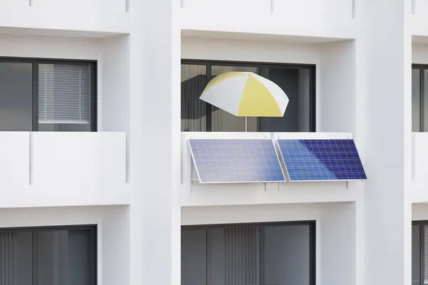 Ein Mehrfamilienhaus. An einem Balkon hängt ein Balkonkraftwerk und steht ein Sonnenschirm.