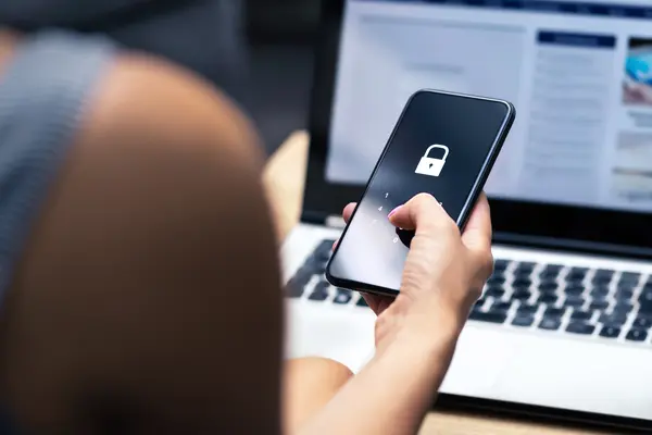 Eine Person sitzt an einem Laptop und hält ein Smartphone in der Hand. Auf dem Smartphone ist ein Verschlüsselungssymbol.