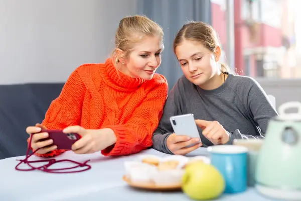 Mutter sitzt mit Tochter am Tisch und schaut auf ihr Smartphone.