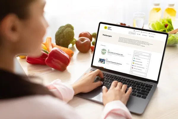 Eine Frau sitzt mit Laptop am Küchentisch und schaut auf eine Internetseite auf dem Bildschirm.