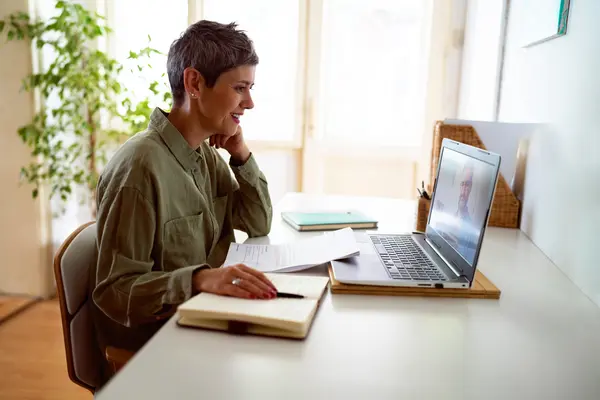 Frau sitzt am Tisch und schaut lächelnd auf den Bildschirm ihres Laptops.