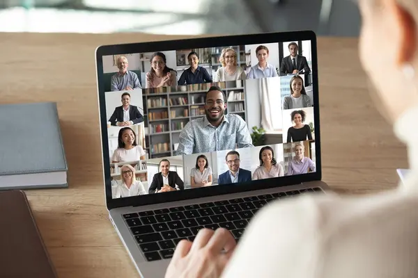 Jemand sitzt vor einem Laptop und hält eine digitale Konferenz ab. Auf dem Bildschirm sind viele Menschen zu sehen, die in die KAmera schauen. 