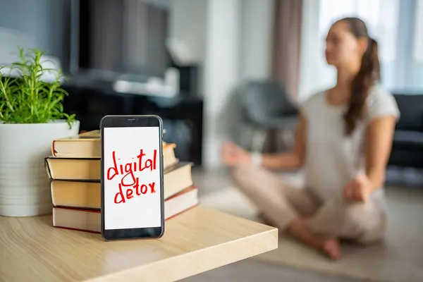 Im Vordergrund liegt ein Handy mit den Wörtern: digital detox auf dem Bildschirm. Im Hintergrund macht eine Frau Yoga