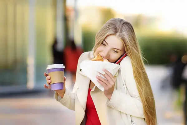 Eine Frau ist unterwegs und beißt in ein Sandwich. Inder anderen Hand hat sei einen Coffee to go in der Hand