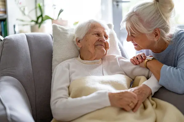 Eine Seniorin Sitzt durch ein Kissen gestützt auf der Couch und lächelt. Eine weitere Frau lehnt sich liebevoll zu ihr rüber und schaut sie an.