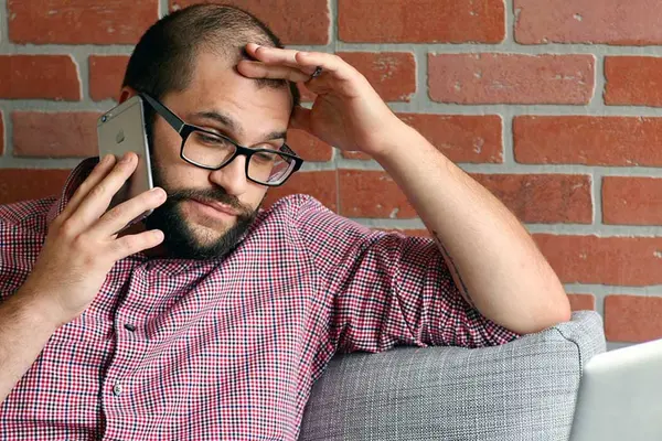 Ein Mann sitzt genervt mit seinem Smartphone am Ohr vor seinem Laptop 