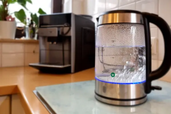 Zu kochen beginnendes Wasser in einem gläsernen Wasserkocher auf der Arbeitsplatte einer Küche, dahinter unscharf ein Kaffeevollautomat.