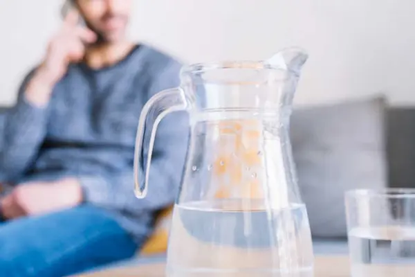Im Hintergrund sitzt ein Mann telefonierend auf dem Sofa, vor ihm eine Karaffe und ein Glas mit Wasser 