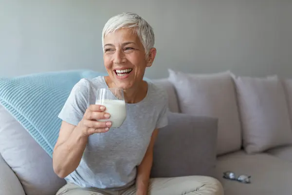Lachende Frau mit grauen Haaren sitzt auf der Couch mit einem Glas Milch in der Hand 