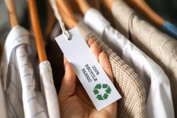 Kleidung auf Kleiderbügel, Hand hält Etikett auf dem steht "100% Recycle Fabric"