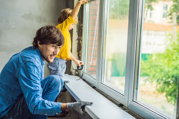 Ein Mann tauscht ein Fenster aus. Er montiert gerade die Fensterbank an oder ab. Ein kleiner Junge unterstützt ihn. 