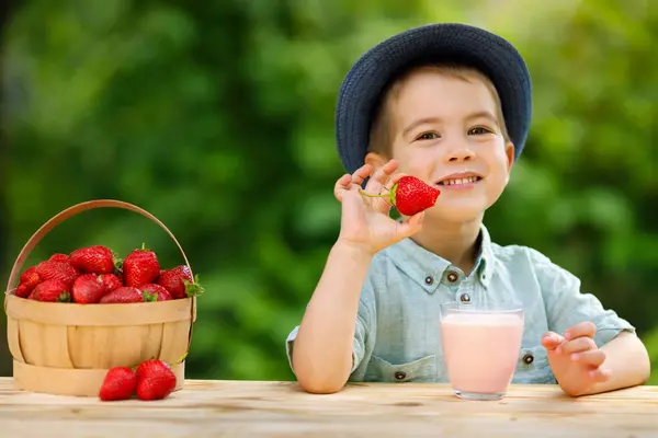 Ein Junge sitzt mit einem Glas Erdbeermilch und einem Korb Erdbeeren am Tisch