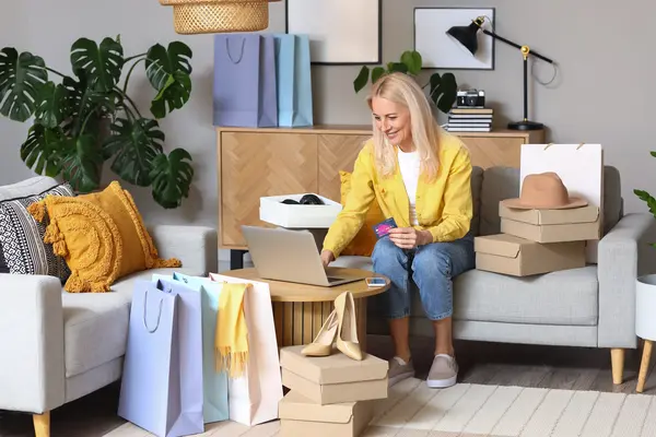 Eine Frau sitzt auf dem Sofa vor ihrem Laptop, in der Hand hält sie eine Kreditkarte. Sie lächelt, um sie herum sind viele Einkaufstüten verteilt, sowie ein Paar Highheels und ein Hut.