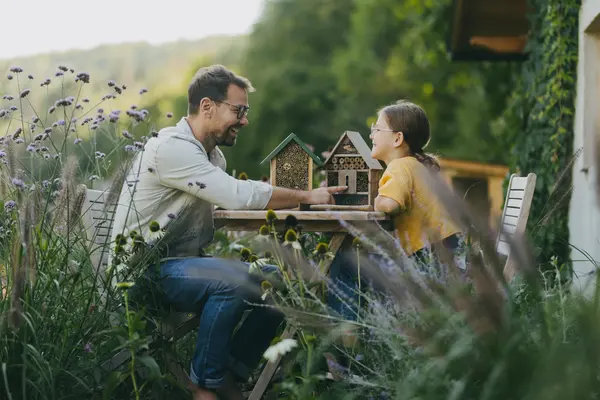 Vater und Tochter sitzen gemeinsam an einem Tisch im Garten umgeben von hohem Gras und Wildblumen und lachen sich an. Auf dem Tisch stehen zwei Insektenhäuser.