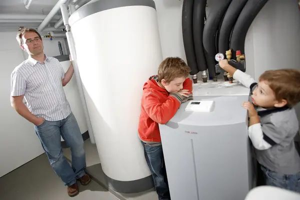 Ein Vater steht im Keller seines Hauses neben seiner Wärmepumpe. Zwei Kinder sind auch anwesend.