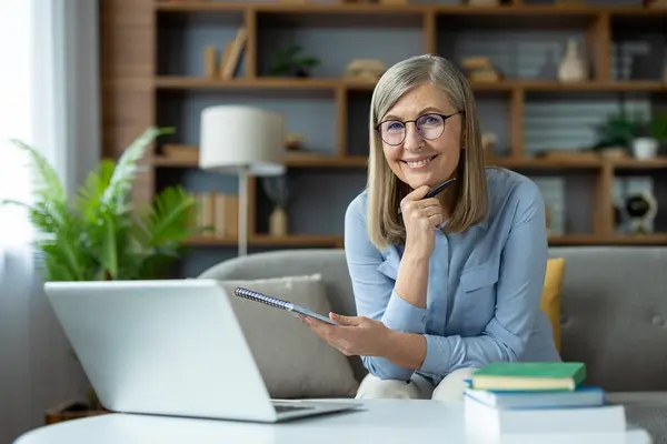 Eine Frau mittleren Alters mit Brille sitzt auf dem Sofa, vor ihr steht ihr Laptop und ein Stapel Bücher. Sie hält ein Notizblock in der einen Hand und einen Kugelschreiber in der anderen. Diese Hand hat sie an ihr Kinn gestützt und lacht freundlich in die Kamera.