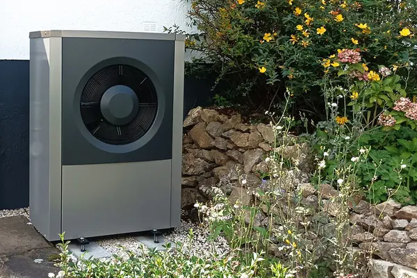 Graue Wärmepumpe vor einem Einfamilienhaus mit Blumen