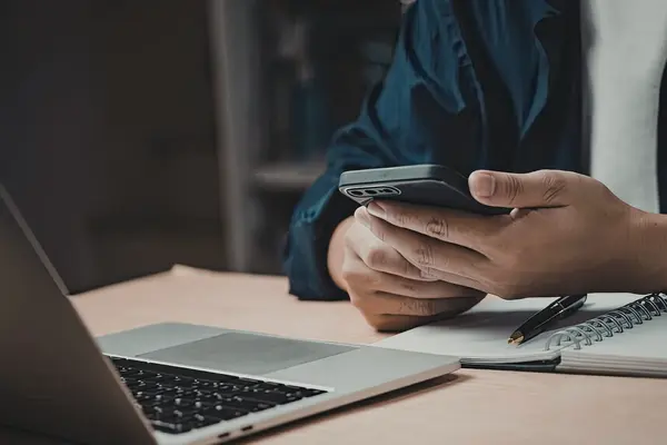 Person am Schreibtisch mit Handy in der Hand vor Laptop