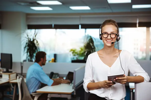Eine Frau mit Headset steht in einem Büro, hinter ihr sitzt ein Mann am PC