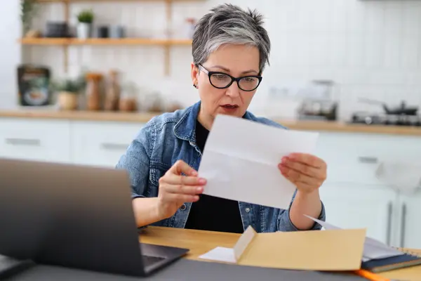 Eine Frau sitzt an einem Tisch mit Laptop und blickt erschrocken auf einen Brief.