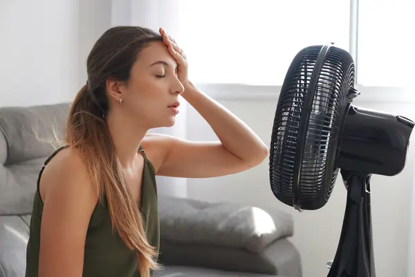 Erschöpfte Frau sitzt bei Hitze vor einem Ventilator im Wohnzimmer.