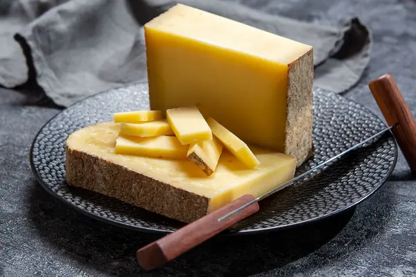 Zwei große Stücke und mehrere kleine Stücke Bergkäse liegen auf einem schwarzen Teller, daneben ein Käsemesser