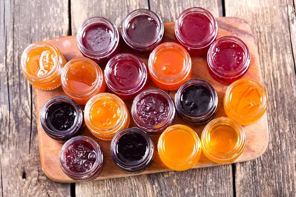 Verschiedene geöffnete Gläser mit Marmelade stehen auf einem Tablett. Man blickt von oben auf die geöffneten Gläser und sieht gelbe, orangefarbene, rote und dunkelrote Marmeladen.
