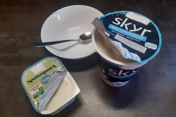 Eine Packung Quark und ein Becher Skyr stehen auf einem Tisch neben einer weißen Schüssel, in der ein kleiner Löffel liegt.