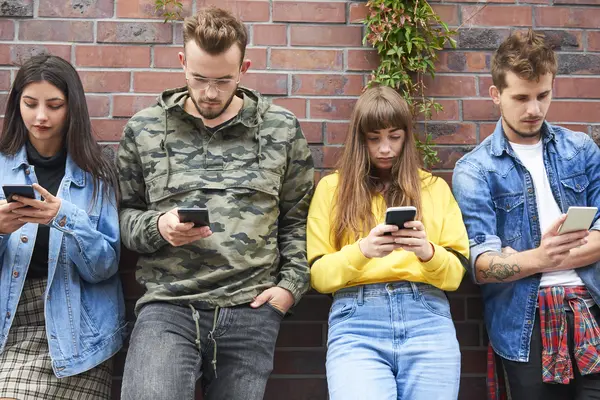Vier junge Erwachsene stehen an eine Mauer gelehnt und schauen in ihr Handy.