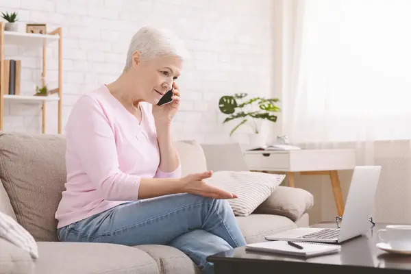 Eine ältere Frau sitzt zu Hause auf dem Sofa und telefoniert mit dem Smartphone. Vor ihr steht ein geöffneter Laptop auf dem Couchtisch. Mit der freien Hand gestikuliert sie und wirkt besorgt oder ratlos.