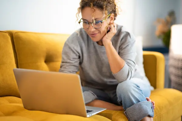 Eine Frau sitzt auf einem gelben Sofa vor einem Laptop 