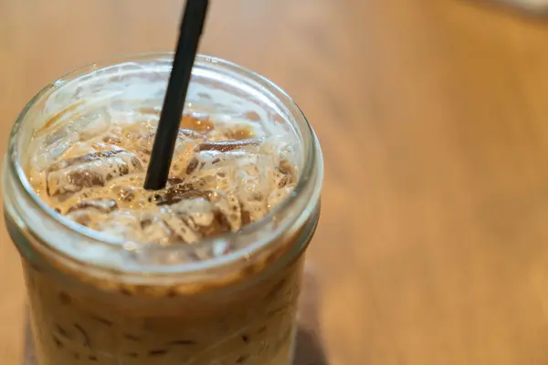 Ein Glas mit Kaffee und Eiswürfeln auf einer Holzoberfläche. In der Mitte steckt ein Strohhalm.