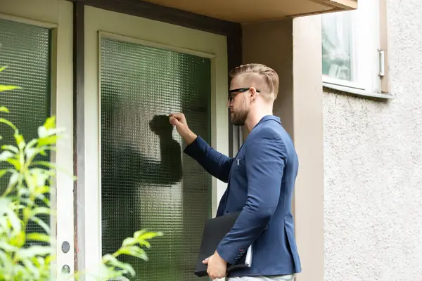 Vertreter in blauem Anzug und Brille klopft an der Türe eines Hauses