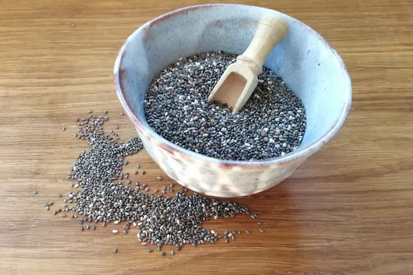 Eine kleine Schüssel mit Chiasamen steht auf einem Holztisch. In der Schüssel steckt ein kleiner Holzlöffel im Chiasamen.