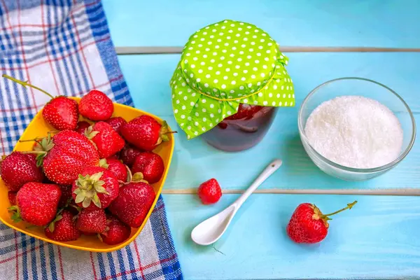 Auf einem Tisch stehen eine Schüssel mit Erdbeeren, ein Glas Marmeladen und eine Schüssel mit Gelierzucker.