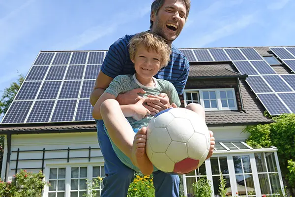 Vater spielt mit Sohn Ball. Im Hintergrund ist Haus mit Solaranlage zu sehen.